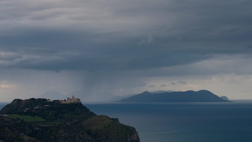 Un santuario illuminato sulla cima di un promontorio sul mare, sotto un temporale, con le isole sullo sfondo