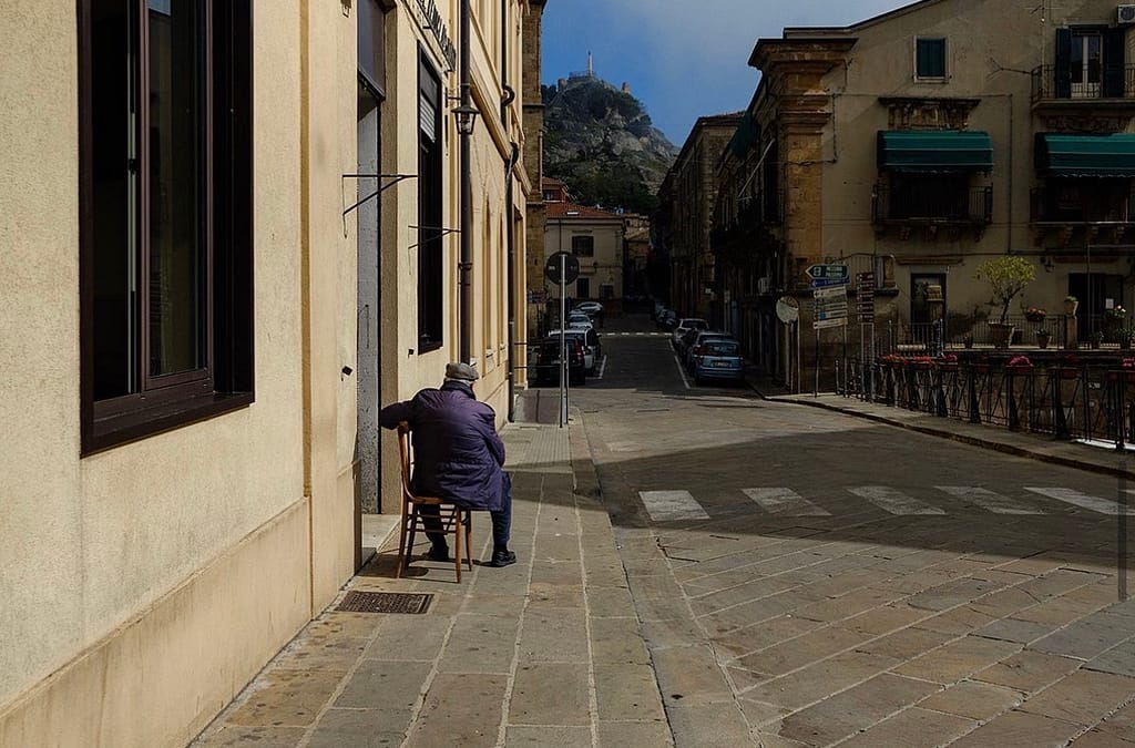 Anziano seduto di spalle su una sedia in una strada di paese, con una rocca sullo sfondo.