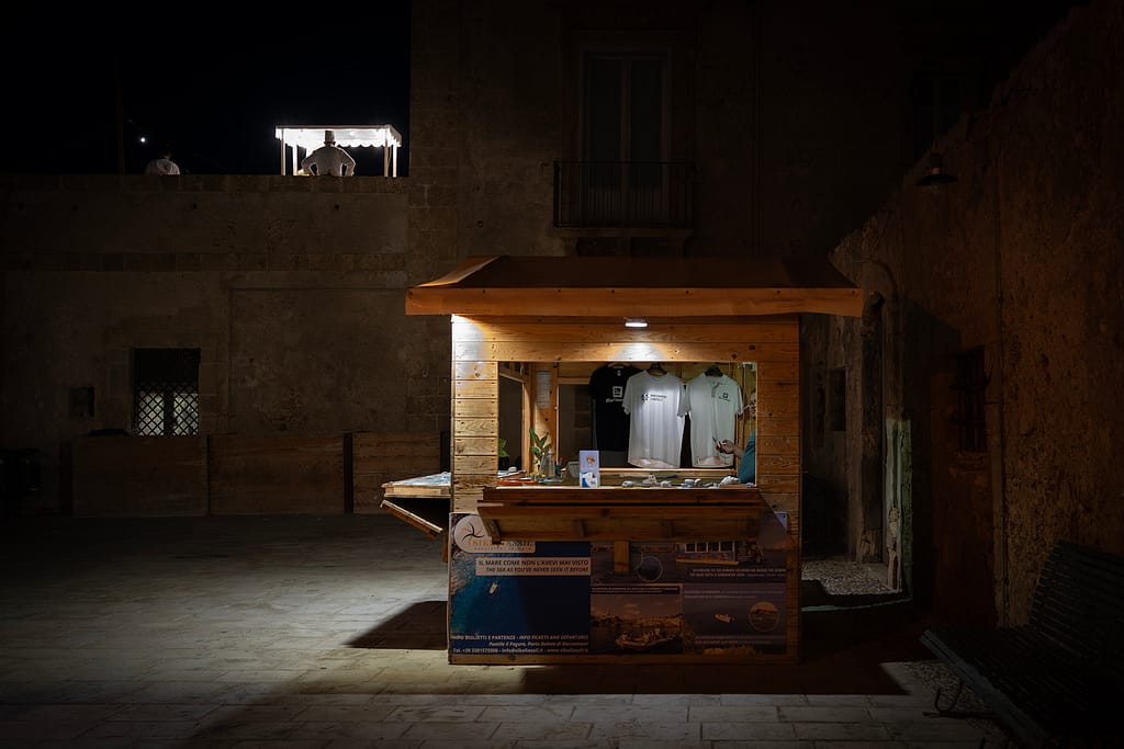 Chiosco di legno illuminato in una piazza buia di Marzamemi, con sopra una terrazza dove un cuoco di spalle lavora sotto un gazebo luminoso.