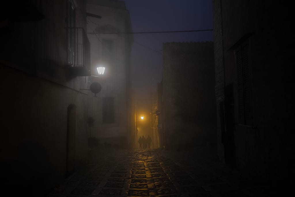 Vicolo di Erice di notte nella nebbia con lampioni e tre persone in fondo