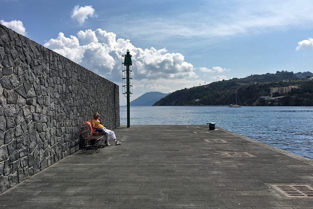 Uomo seduto su una panchina sul molo di Lipari, accanto a un muro di pietra lavica e al mare