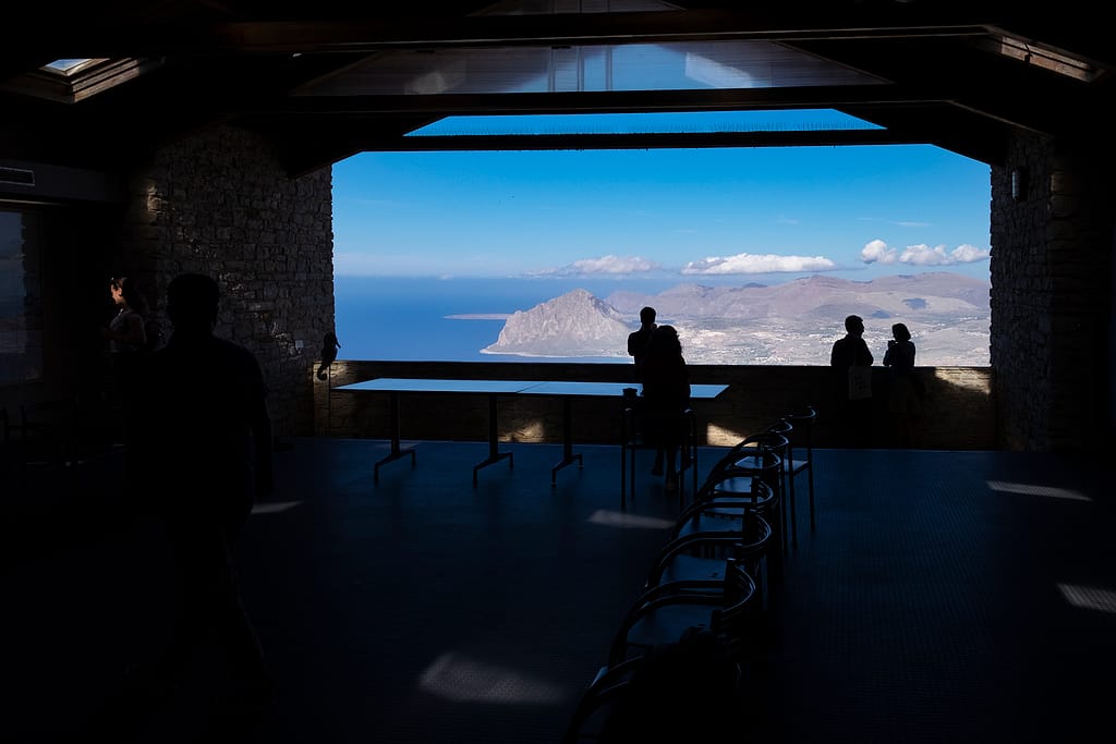 Grande apertura della sala buia dell'osservatorio di Erice che inquadra il mare e il Monte Cofano, con alcune persone in controluce affacciate al parapetto.
