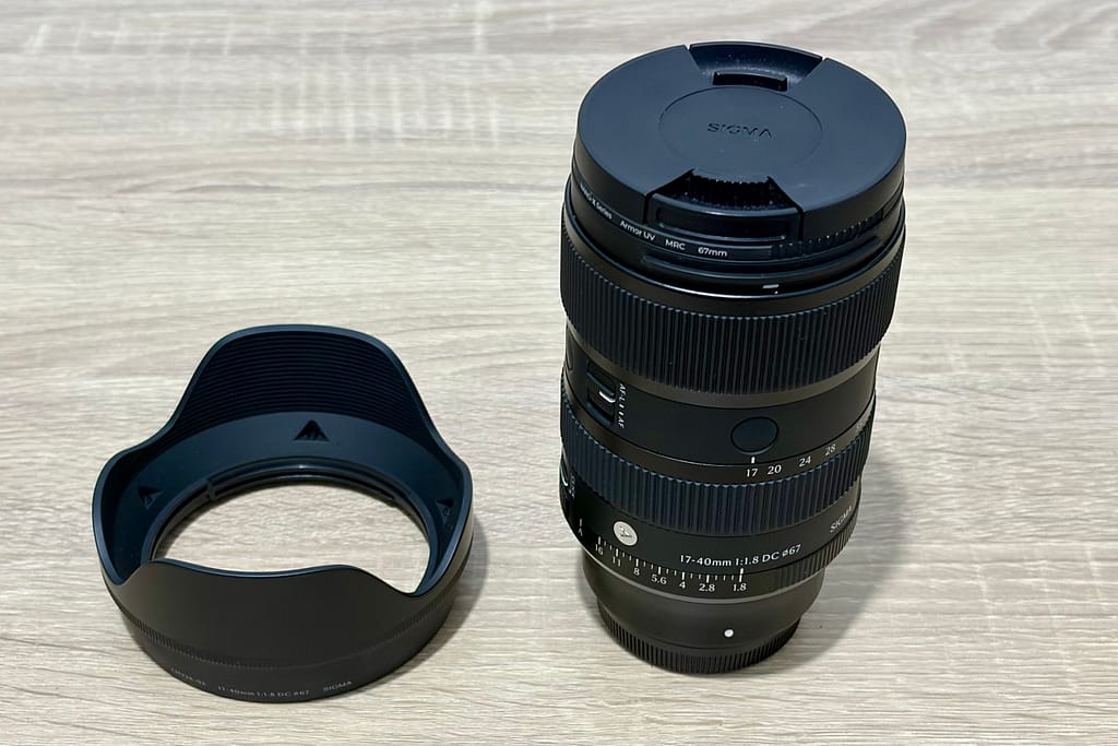 Obiettivo Sigma 17-40mm f/1.8 DC Art appoggiato su un tavolo di legno chiaro, accanto al suo paraluce.
