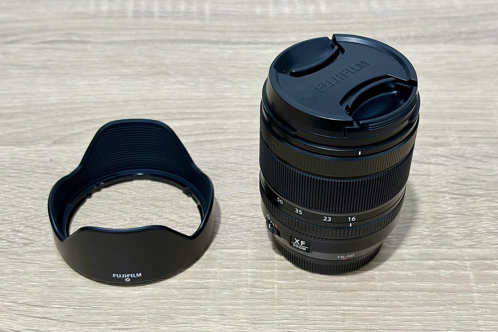 Obiettivo Fujifilm XF 16-50mm f/2.8-4.8 con il tappo, appoggiato su un tavolo di legno chiaro accanto al suo paraluce.