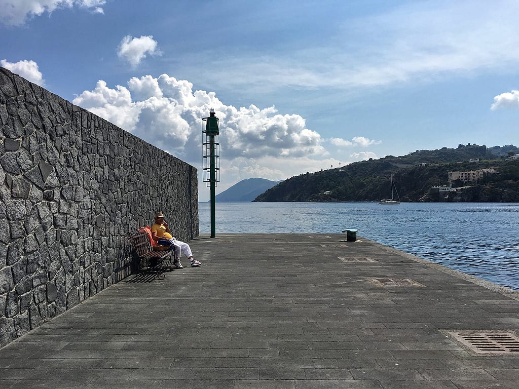 Uomo seduto su una panchina sul molo di Lipari, accanto a un muro di pietra lavica e al mare
