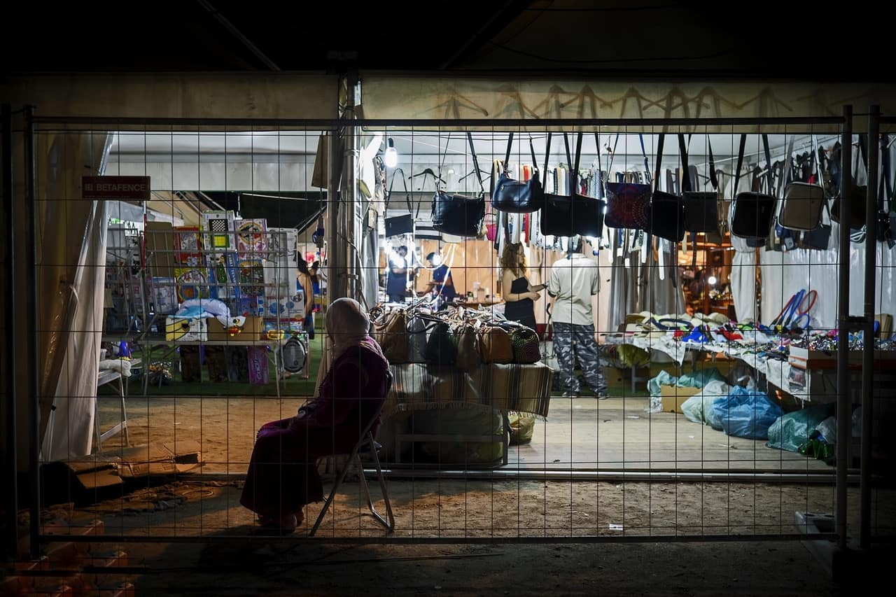 Donna con il velo seduta di notte davanti a un mercato illuminato, vista attraverso una rete.