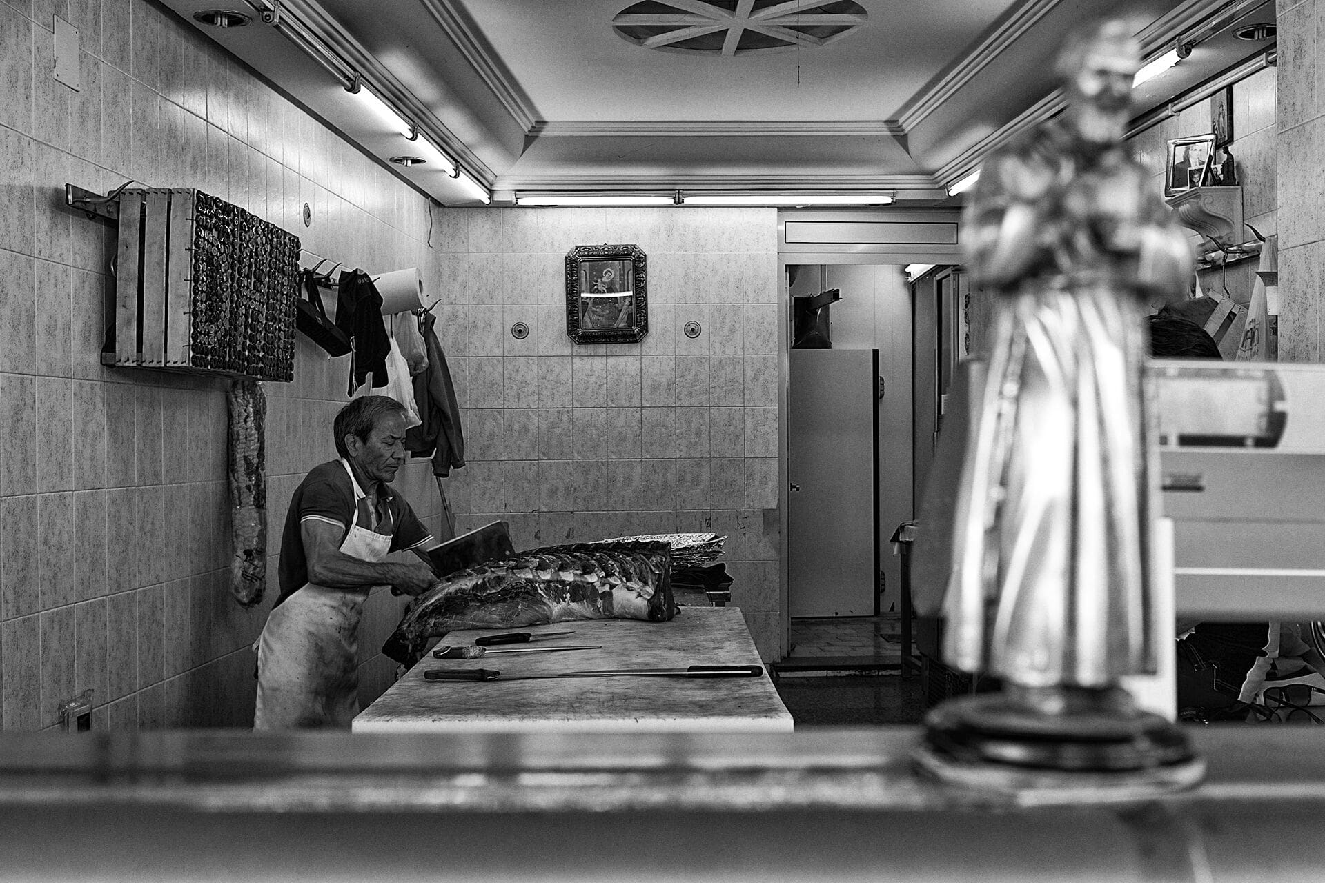 In bianco e nero, un macellaio al lavoro nella sua bottega a Palermo, con una statuetta in primo piano