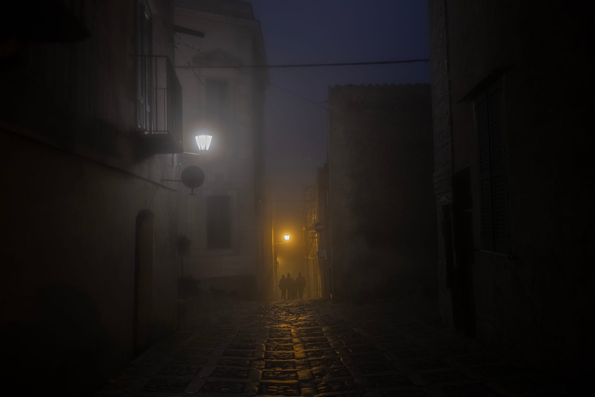 Vicolo di Erice di notte nella nebbia, con due lampioni accesi e tre persone in controluce in fondo