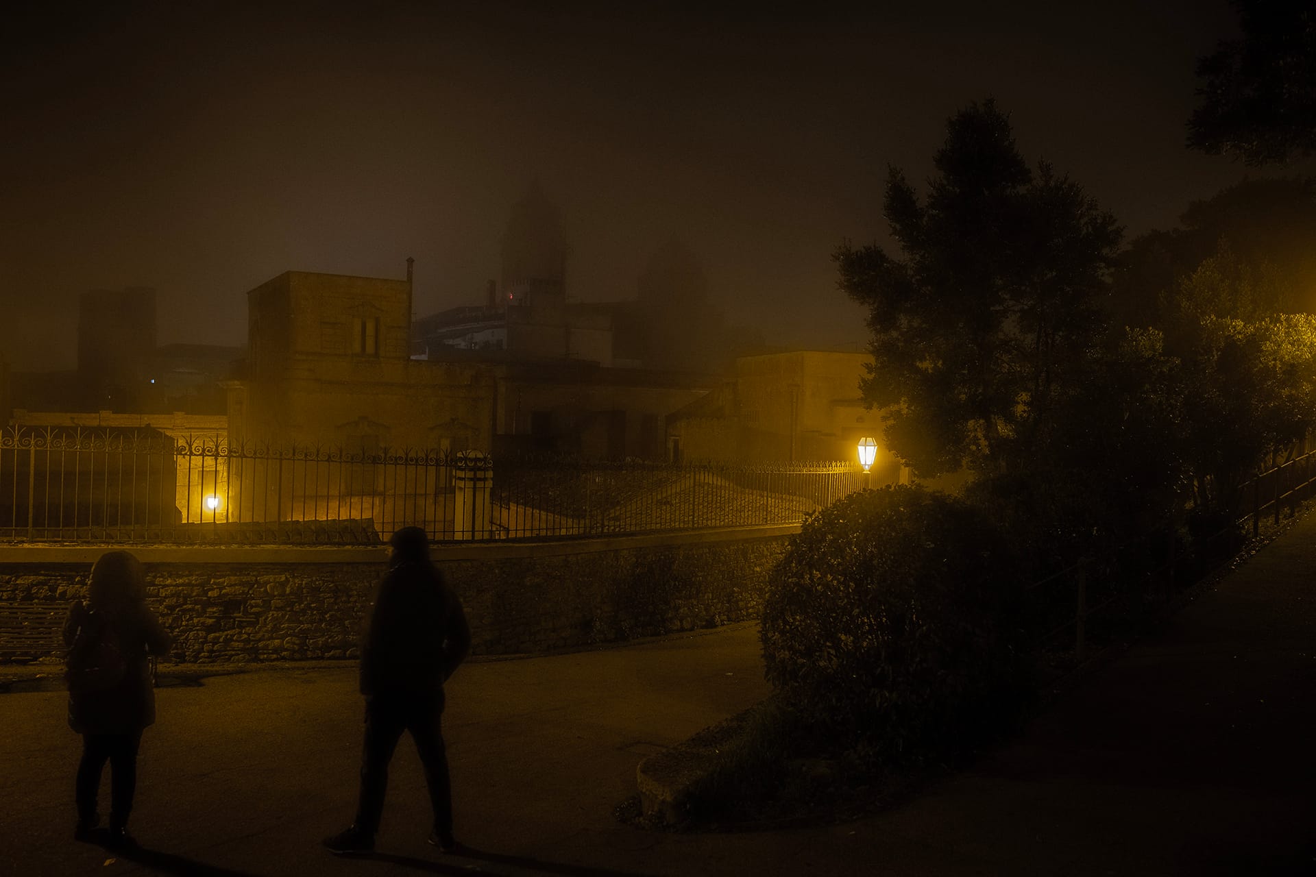 Due persone in un piazzale di Erice di notte, con la nebbia e la luce gialla dei lampioni sugli edifici