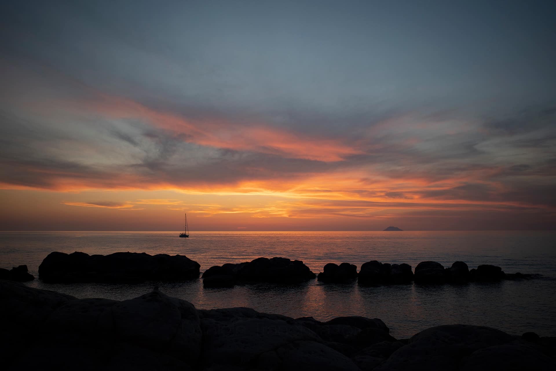 Tramonto sul Tirreno con una barca a vela e un’isola all’orizzonte, scogli in controluce in primo piano