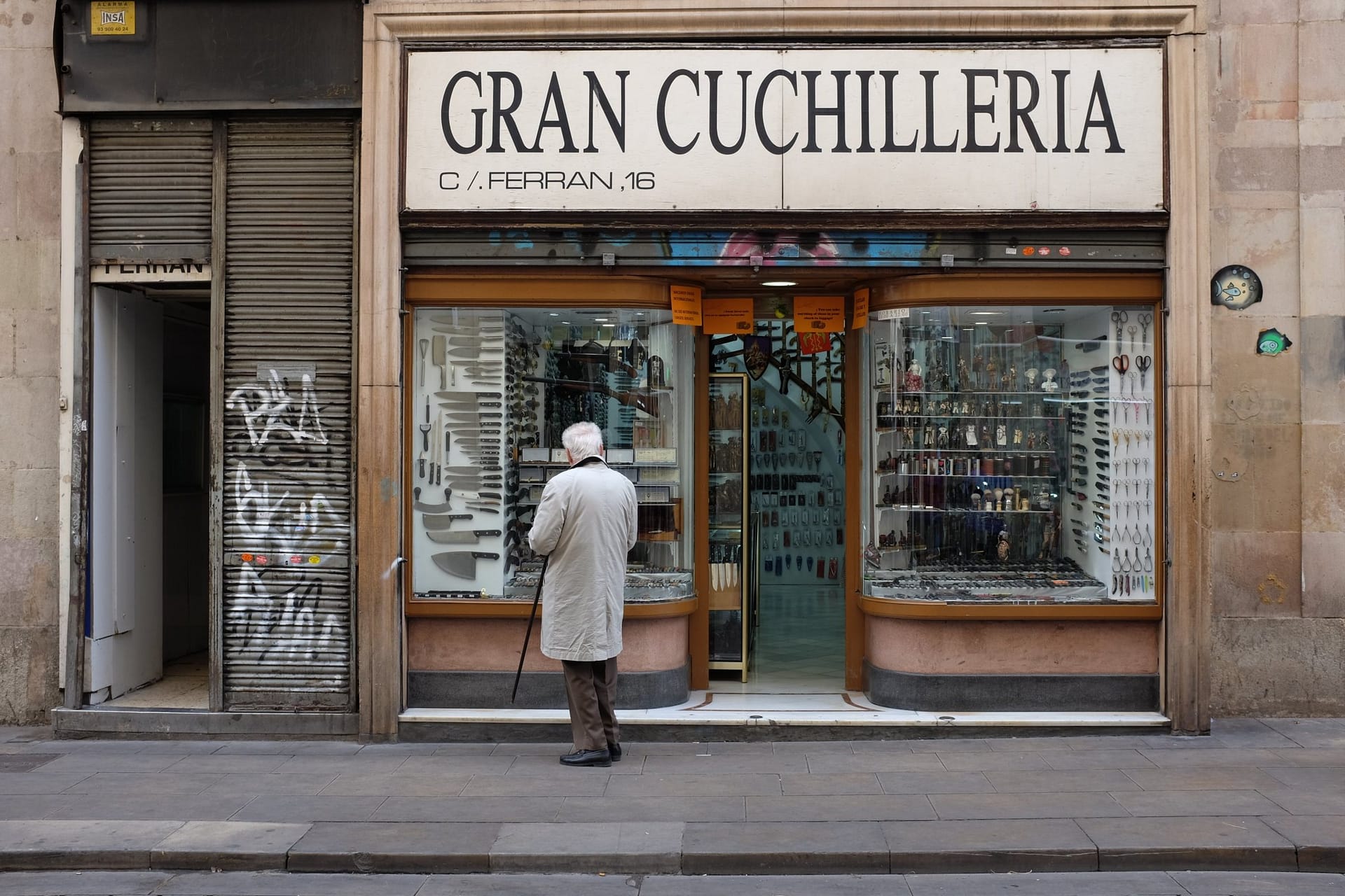 Un signore anziano con il bastone, di spalle, guarda la vetrina di una vecchia coltelleria a Barcellona