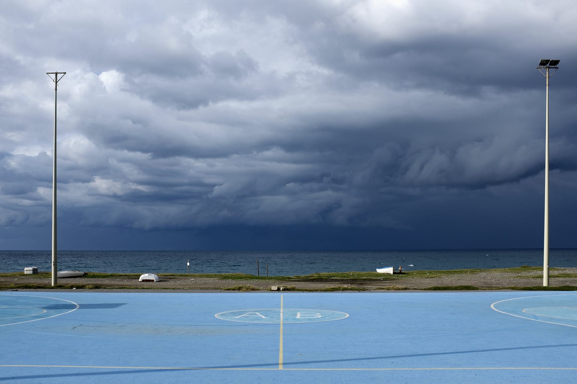 Un campo da gioco azzurro in riva al mare, con due pali della luce e un temporale scuro che arriva dal mare