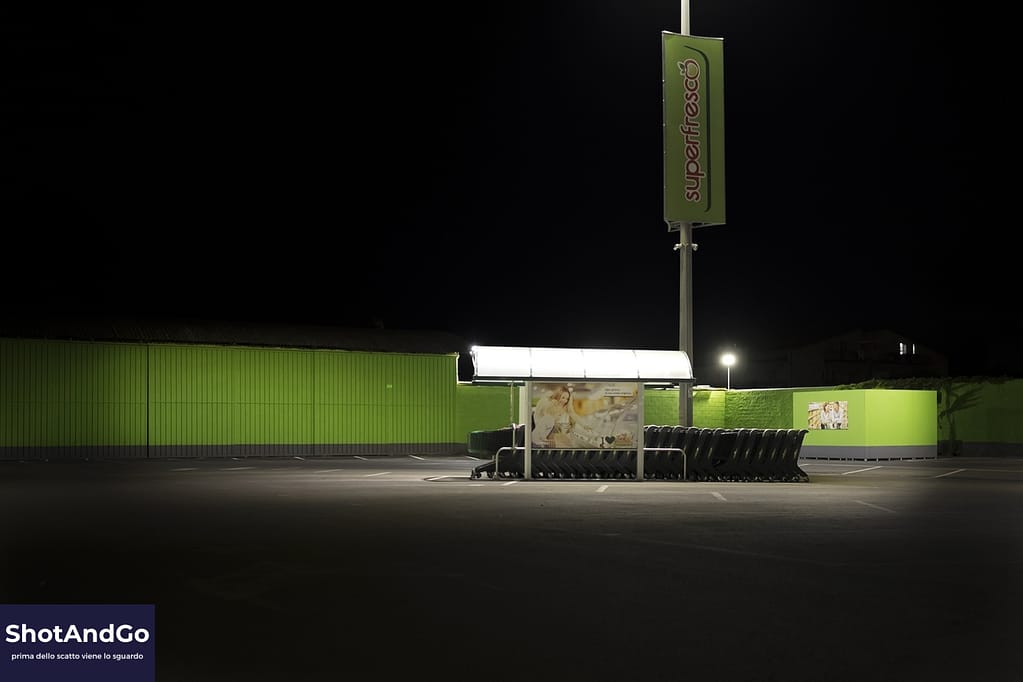 Parcheggio di un supermercato illuminato di notte con un edificio isolato e ampi spazi vuoti.