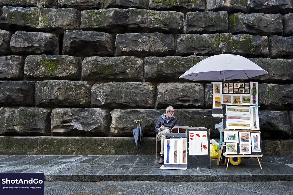 Artista di strada espone le proprie opere davanti a un muro di pietra, simbolo di una fotografia fatta di attesa e consapevolezza.