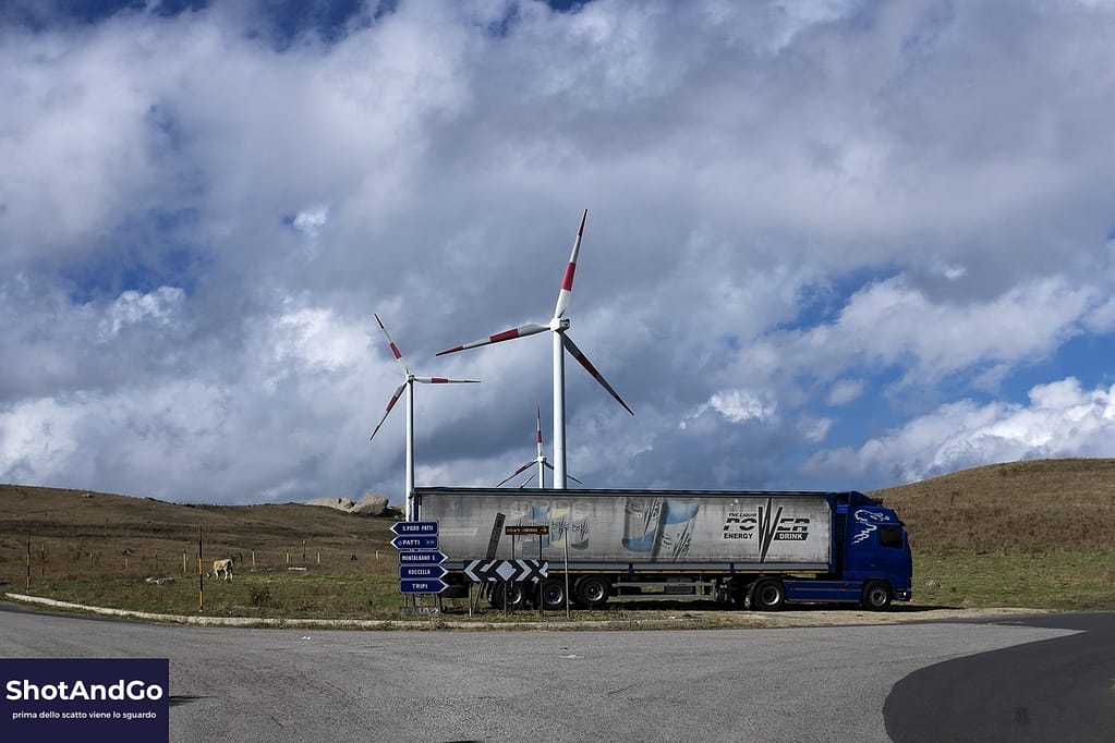 Perché fotografare oggi (quando siamo già sommersi di immagini) Camion che attraversa un paesaggio con pale eoliche sotto un cielo nuvoloso, simbolo della fotografia contemporanea