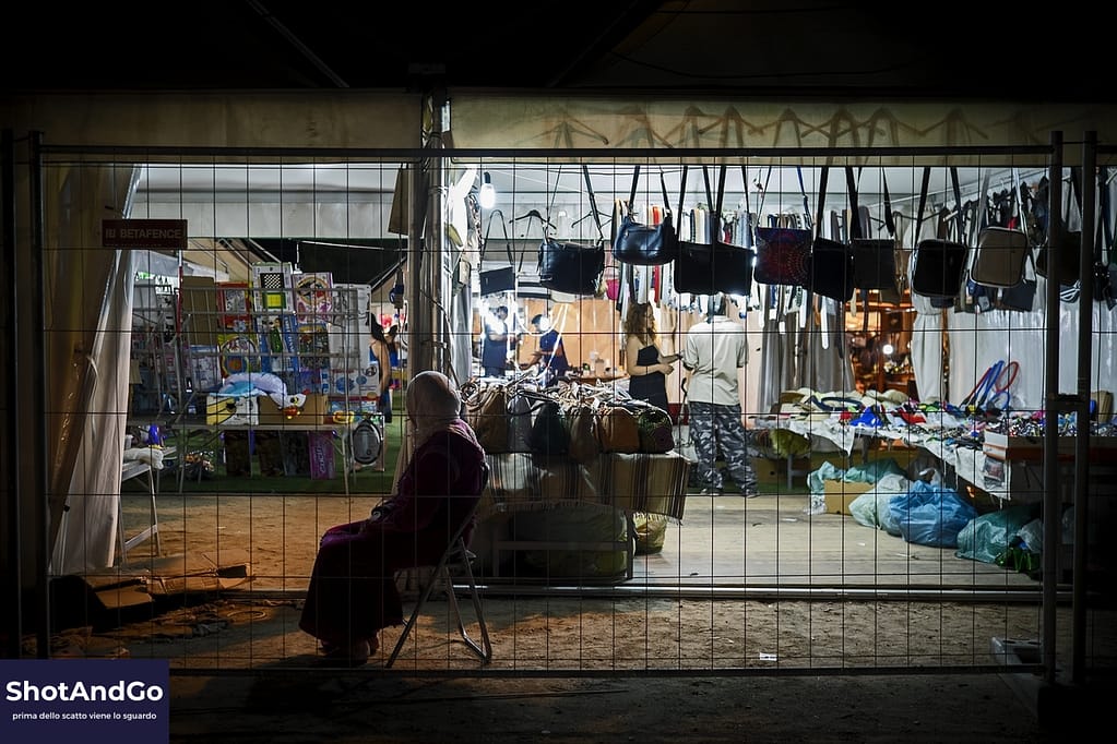 Donna seduta davanti a una bancarella di un mercato notturno osservata da lontano, esempio di street photography rispettosa e narrativa.