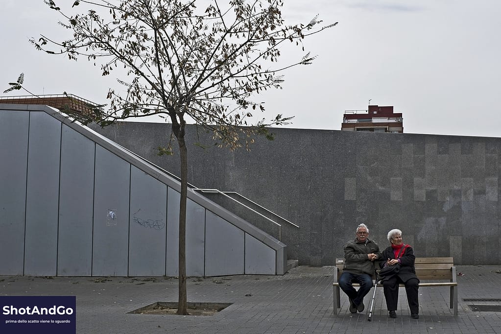 Due persone anziane sedute su una panchina in uno spazio urbano, esempio di presenza e osservazione nella street photography.