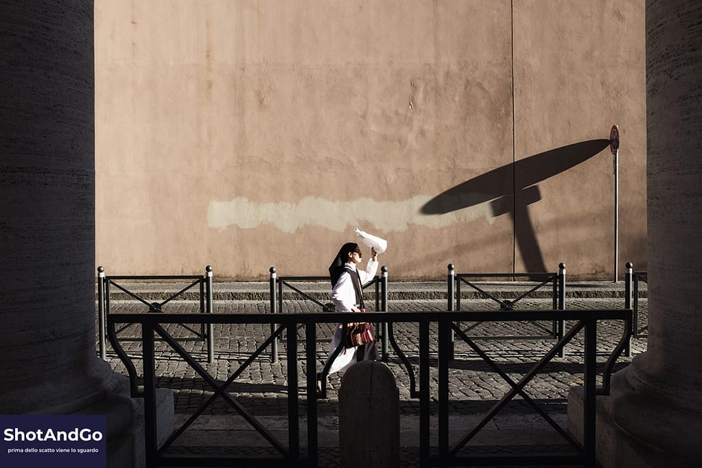 Una donna cammina lungo una strada illuminata dal sole mentre la sua ombra si proietta su un muro, esempio di osservazione nella street photography.
