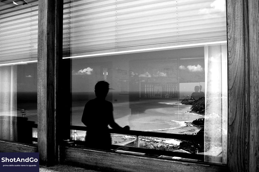 Silhouette di una persona riflessa in una finestra affacciata sul mare, simbolo di un momento di pausa e riflessione nel percorso fotografico.